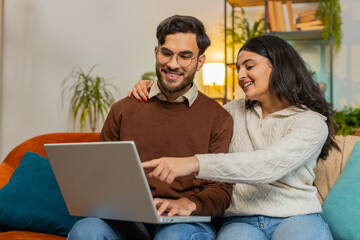 Cheerful diverse couple in casual clothes discussing while using laptop on sofa in living room at home. Happy Hispanic family planning together pointing showing booking online tickets in apartment.