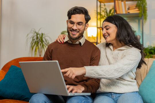 Cheerful diverse couple in casual clothes discussing while using laptop on sofa in living room at home. Happy Hispanic family planning together pointing showing booking online tickets in apartment.