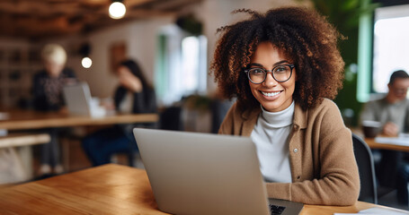 Smiling young woman with curly hair and glasses working on a laptop in a modern, collaborative office setting.
