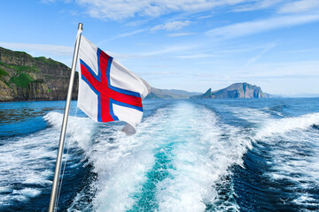 View of the Atlantic ocean and Faroe Islands from a boat sailing from the city of Sorvagur to the island of Mykines. Flag of Faroe islands waving against a cloudy sky. Landscape photography