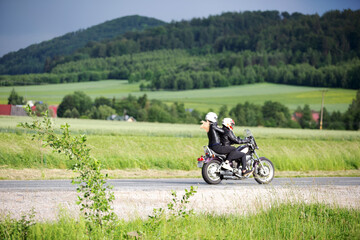 Motocykl na drodze z pasażerką, zielone pola, dachy domu, lasy na wzgórzu. © Stanisław Błachowicz