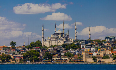 Naklejka premium The Blue Mosque stands tall against a backdrop of blue sky and fluffy white clouds. A turquoise body of water extends towards the viewer.