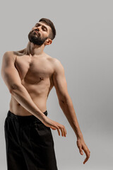 Shirtless man with beard, wearing black pants, in creative pose that highlights his athleticism against warm grey background. Concept of man's fashion and style, beauty and youth, masculinity.