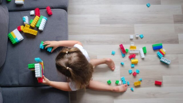 Top view of pretty little caucasian girl playing plastic colorful cubes toy alone, sitting on warm wooden floor with underfloor heating, cute adorable toddler child having fun on couch at home