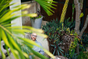 male casual man with the beard in a grey sweetshot in the yard of a tropical villa resting in the evening under a palm tree 