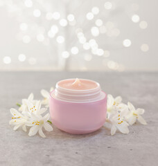 Cream in jar and beautiful jasmine flowers on grey table against light background with blurred lights, closeup