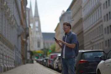 A man using smartphone wearing white sunglasses and gray backpack on the street in Vienna on a hot sunny summer day. Tourism and sightseeing concept