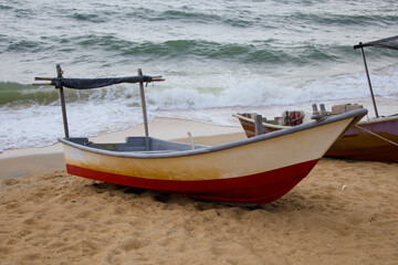 Naklejka premium A vibrant red and white traditional fishing boat rests on the sandy beach of Pantai Teluk Lipat, Dungun, Terengganu. Equipped with wooden oars, it appears ready for a day at sea.