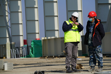 Two multiracial construction workers or engineers walking side by side through a construction site, conversing.                        