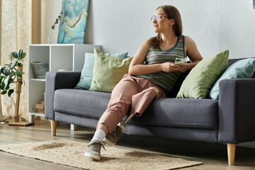 Obraz premium A young woman with a prosthetic leg sits on a sofa in her living room, holding her phone and looking out the window.