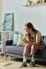 Obraz premium A young woman with a prosthetic leg smiles while looking at her phone while sitting on a couch in her living room.
