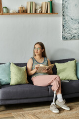 Obraz premium A young woman with a prosthetic leg sits on a sofa in her living room, reading a book and gazing thoughtfully out of the window.