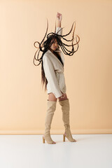 A young woman in stylish attire poses on a beige backdrop. Her long, dark braids flow freely around her.
