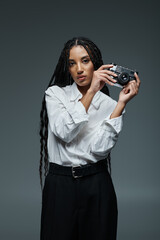 A young woman with long braids poses confidently while holding a camera in front of a grey background.