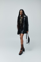 A young woman in a black leather jacket and skirt poses confidently against a grey background, her...