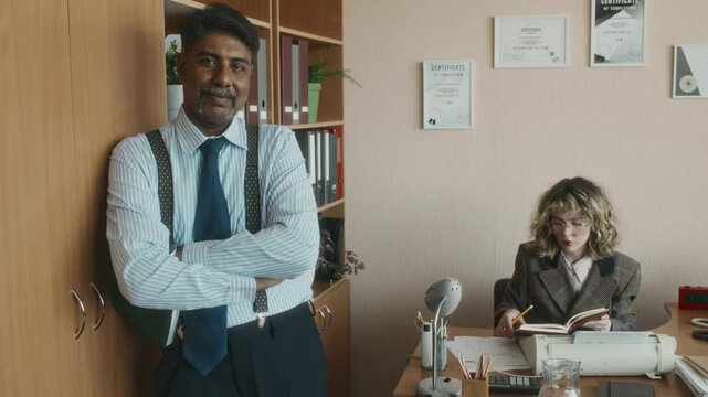 Portrait of mature diverse male boss in striped shirt, tie and suspenders posing with notepad in 90s style retro office, smiling and looking at camera, female secretary working in background