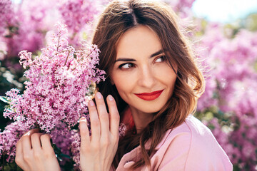 Young beautiful smiling woman. Sexy carefree model posing in the street. Positive model near blooming flower lilac bush beaming. Summertime, pink colours. Closeup beauty portrait, red lips