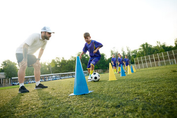 Young boy, child in motion, dribbling soccer ball around cones and receiving guidance from coach....