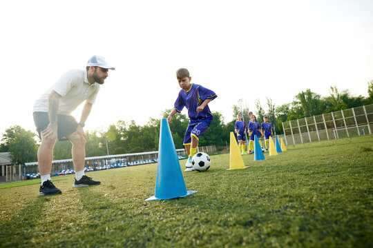 Young boy, child in motion, dribbling soccer ball around cones and receiving guidance from coach. Outdoor stadium game. Concept of sport, childhood, education, achievement, active lifestyle