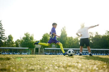 Young boy, soccer player in motion, taking powerful kick while running on filed, coach providing guidance and comments. Concept of sport, childhood, education, achievement, active lifestyle