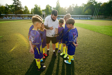 Boys, children in vibrant uniforms gathering around man, coach and listening to game strategy using tablet. Outdoor stadium. Concept of sport, childhood, education, achievement, active lifestyle