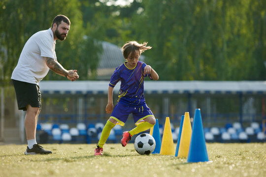 Young soccer player navigating ball through cones, while coach providing instruction and support. Outdoor football game. Concept of sport, childhood, education, achievement, active lifestyle