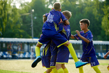 Obraz premium Dynamic moment of victory. Boys, friends in blue uniform, soccer players in motion, jumping and hugging, celebrating winning game. Concept of sport, childhood, education, achievement, active lifestyle