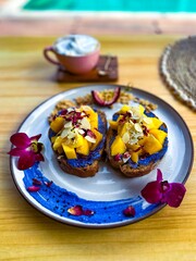 Gourmet toast topped with fresh mango chunks, blue spirulina spread, and almond flakes, garnished with edible flowers, presented on a ceramic plate with a cup of coffee in the background, perfect for