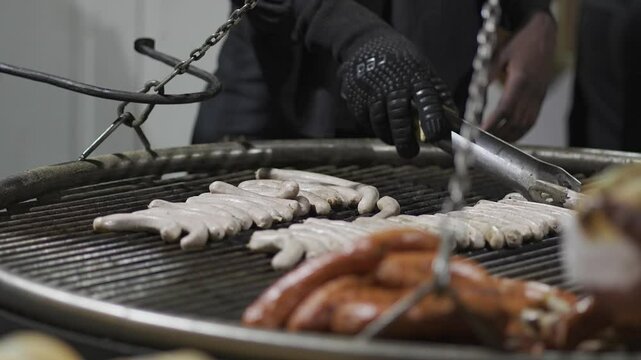 Chefs hands turn sausages on metal grill suspended on chains over fire at Christmas market in Germany in winter. Sausage barbecue of different types in evening at Christmas Fair. Bayerische Wurste. 
