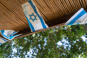 Israeli flags hanging on a wooden pergola with bamboo roofing, captured in an outdoor setting. The...