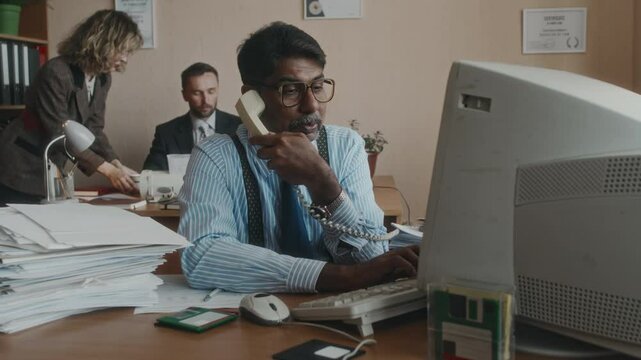 Medium shot of mature Biracial clerical worker with moustache and glasses, wearing formalwear, working on computer and picking up landline phone, sitting next to ream of papers in retro office