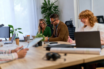 A group of four colleagues having a casual meeting in a modern office. They are sitting around a wooden table with laptops, tablets, and coffee cups.