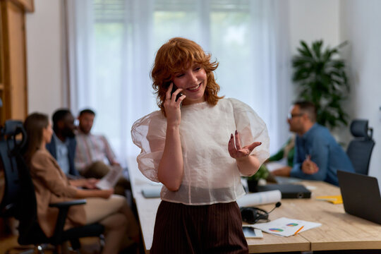 A woman with curly red hair is smiling while talking on the phone in a modern office setting. She is leaning against a desk with papers and a plant on it.