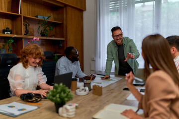 A group of people having a meeting in a conference room. One person is standing and speaking while others are seated and listening. Laptops, notebooks, and coffee cups are on the table.