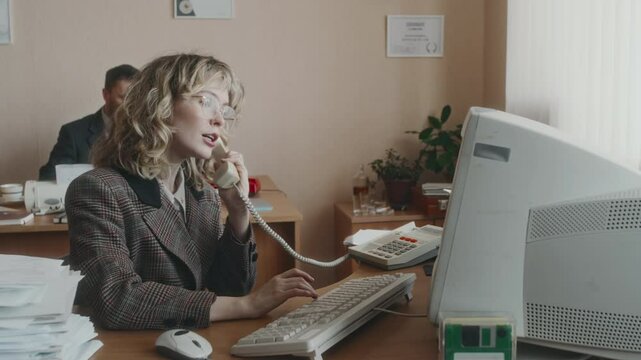 Medium arc shot of fashionable Caucasian secretary with lush blonde hair, wearing checkered jacket and eyewear, working on outdated computer and picking up corded phone, 90s vintage office