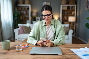 Young business woman using break from working in home office for surfing on social media. Female freelance designer professional using smartphone for text messaging communication.