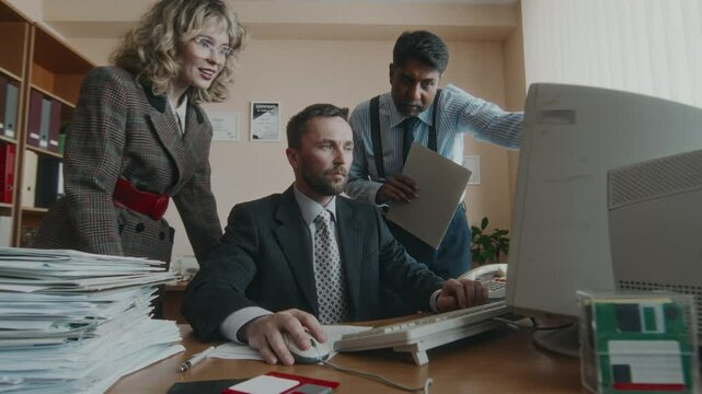 Medium long shot of Caucasian director working on outdated computer and positive diverse subordinates coming up to him, working as team on project in retro office in 90s