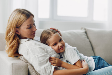 Fototapeta premium Mother and daughter bonding on a cozy living room couch in front of a sunlit window, sharing a warm moment