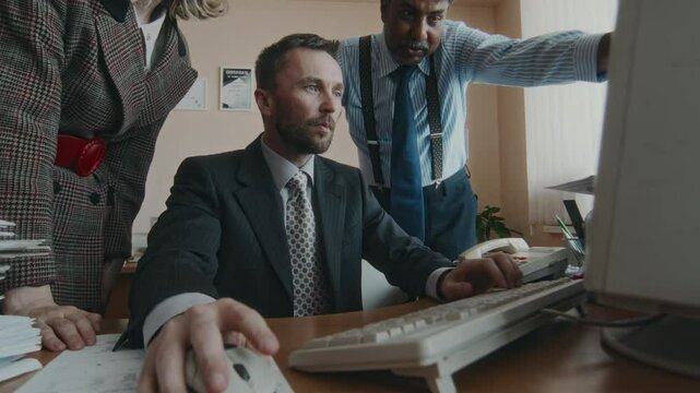 Side shot of good-looking Caucasian businessman working on financial project, typing on outdated computer, male and female subordinates helping him and giving advice on development in 90s retro office