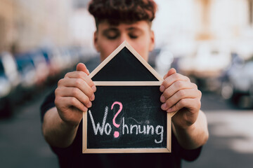 Mann hält Tafel als Haus vor sein Gesicht mit der Schrift Wohnung in rot und weiß