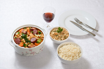 A bowl of chicken cassoulet and spicy breadcrumbs in a bowl, rice in a bowl, a glass of red wine and a plate with cutlery, on a white table.