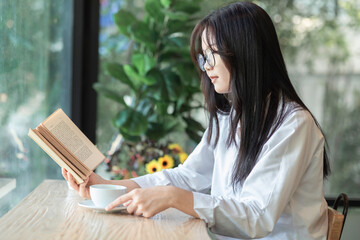 Obraz premium Young asian woman sitting in front of window relaxing in her living room reading book and drinking coffee or tea at home.