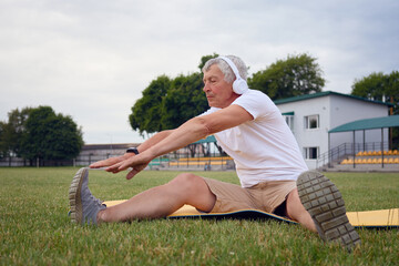 Fototapeta premium Strong Caucasian mature male athlete wearing white T-shirt and wireless headphones on the stadium sitting on yoga mat stretching his legs enjoying urban yoga workout