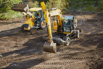Excavator and bulldozer standing on construction site among green trees, initial stage of...