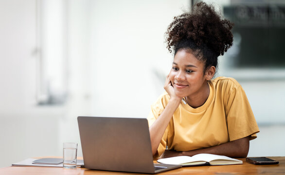 Business And Education Concept. Smiling young black woman student sitting at desk watching on laptop, free copy space. Happy millennial female studying using pc