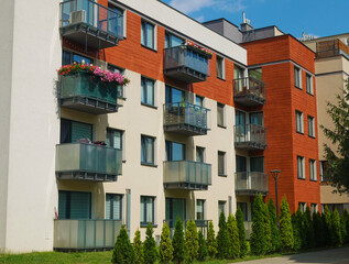 Naklejka premium Modern four-storey residential complex in beige and red colors with glass balconies, near it young thuys planted and green mowed grass 
