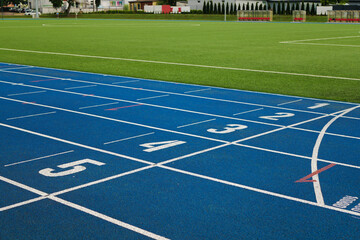 Fototapeta premium Treadmill with blue color and white numbers on it in the vohle of a large soccer field with green artificial grass. There are city buildings in the distance with nice trees growing between the houses.
