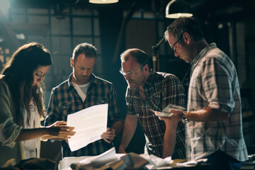 A diverse group of professionals intensely reviews documents under warm industrial lighting, symbolizing teamwork and collaboration in a workshop setting.