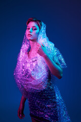 A woman enveloped in bubble wrap stands under vibrant neon lights.