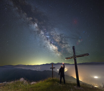 Photograph of Milky Way with person looking at the horizon with light in hand arm raised to the sky next to the crosses of the Ermita del Alba, Quiros, Asturias, Spain.